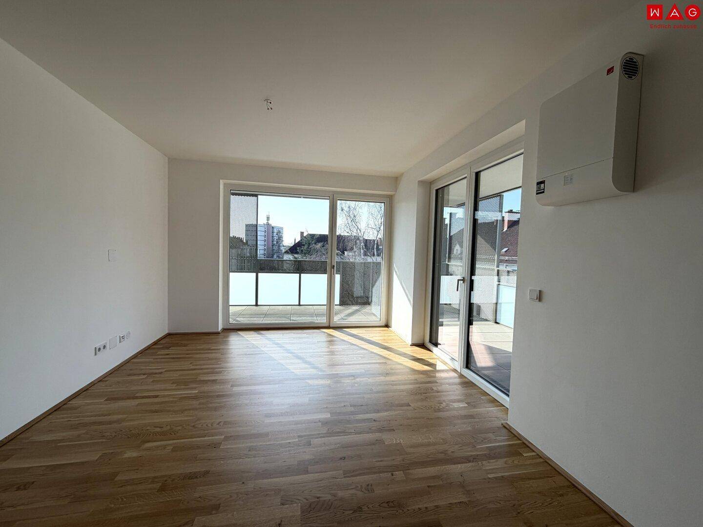 Geräumiges, leeres Wohnzimmer mit Holzboden und großen Fenstern zum Balkon.