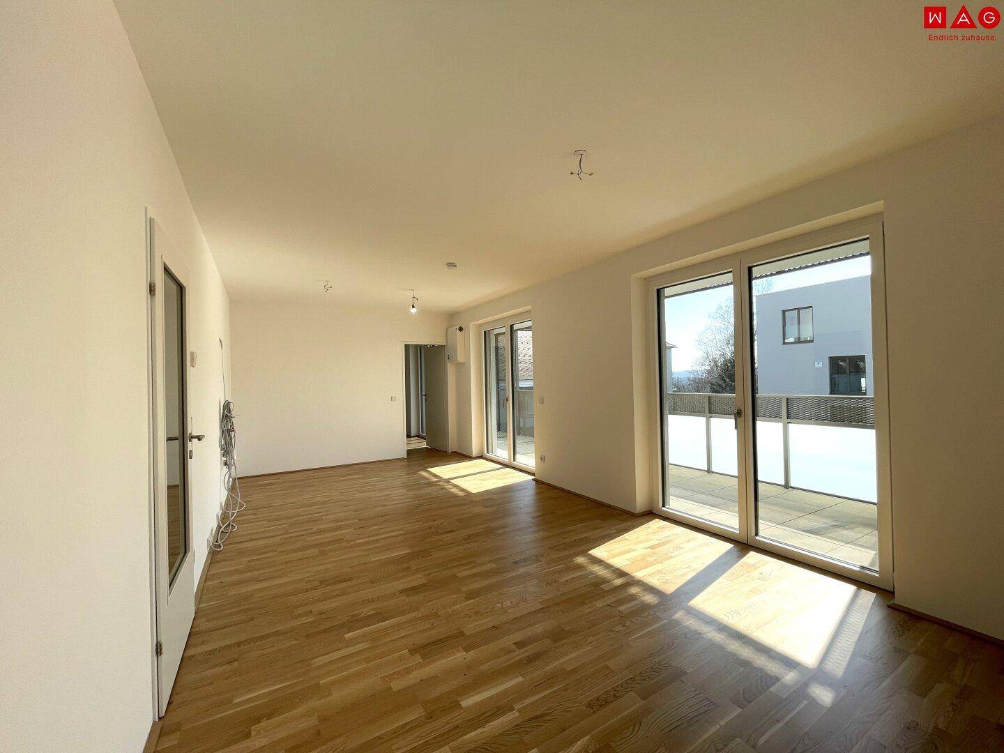 Geräumiges, leeres Wohnzimmer mit Parkettboden und großen Fenstern, die viel Tageslicht hereinlassen.
