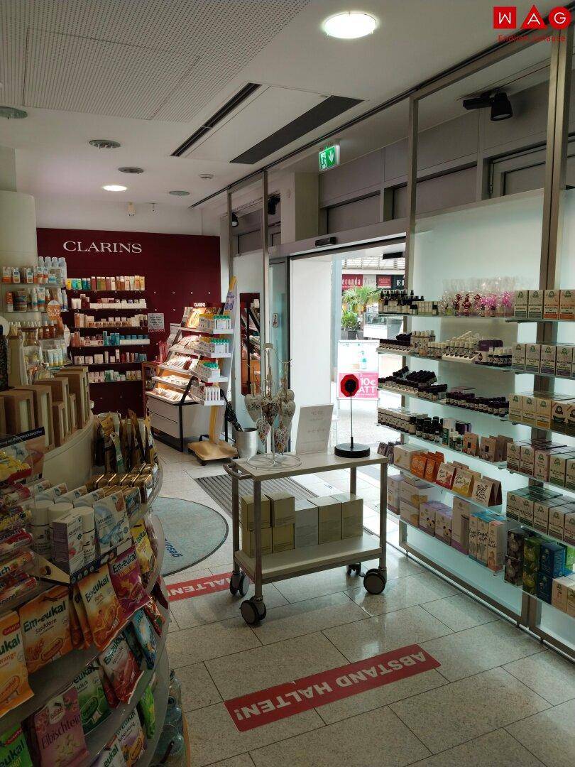 Gang in einer Apotheke mit Produktregalen, einem Rollwagen und Hinweisen auf dem Boden.