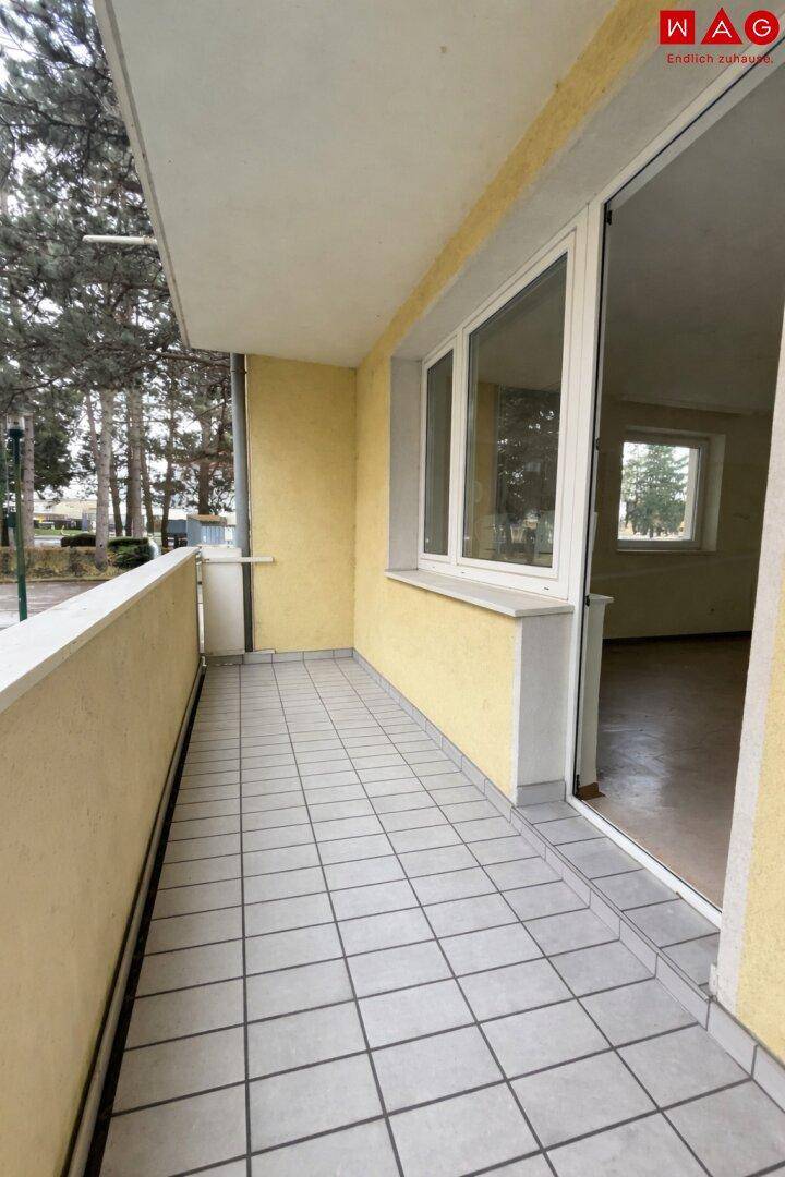 Langer Balkon mit grauen Fliesen und Blick auf Bäume im Hintergrund.