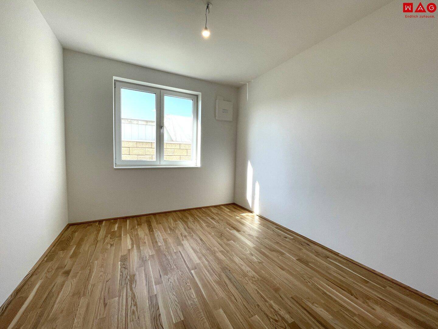 Leeres Schlafzimmer mit Holzboden und einem Fenster, das viel Tageslicht hereinlässt.