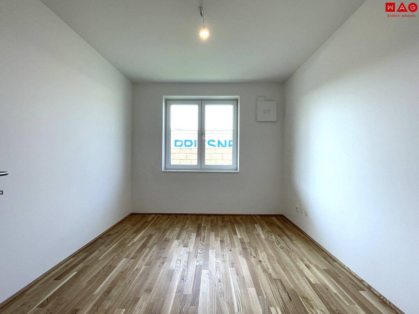 Leeres Schlafzimmer mit Holzboden und einem Fenster, das viel Tageslicht hereinlässt.