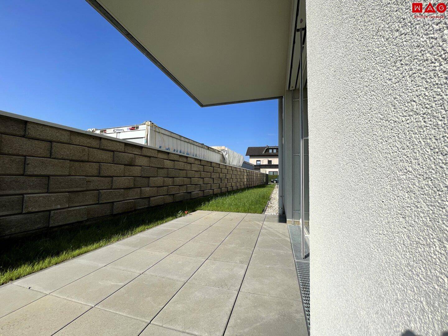 Langer, gepflasterter Terrassenbereich entlang der Gebäudeseite mit angrenzender Grünfläche.