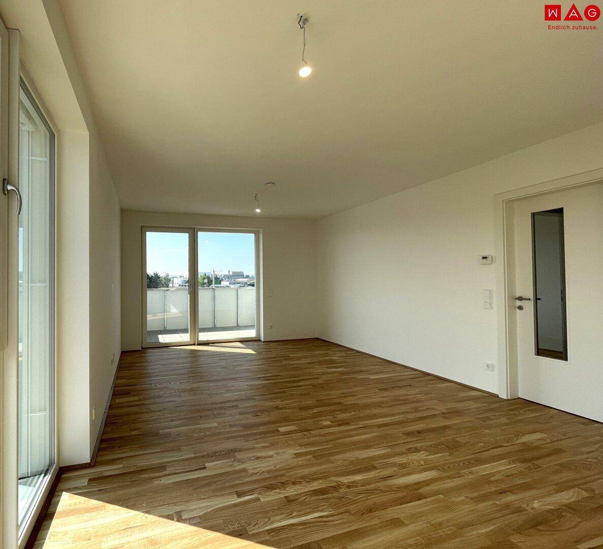 Geräumiges, leeres Wohnzimmer mit großen Fenstern, Holzboden und Zugang zu einem Balkon.