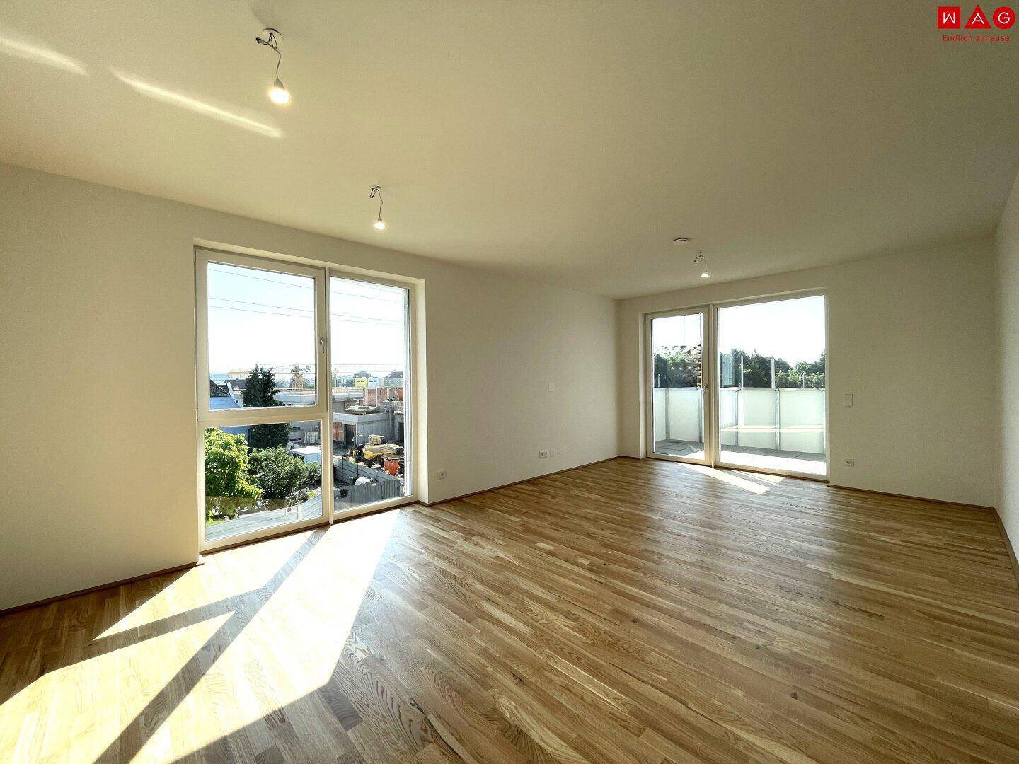 Heller, leerer Raum mit großen Fenstern, Holzboden und Zugang zu einem sonnigen Balkon.