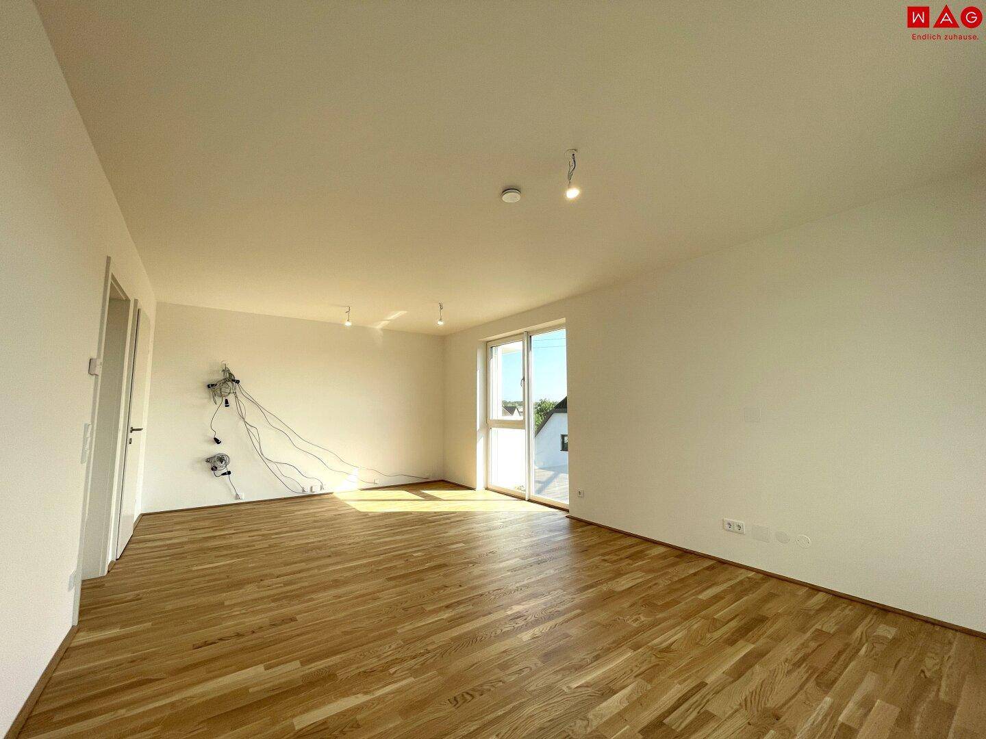Heller, leerer Raum mit Holzboden, großen Fenstern und Zugang zu einem Balkon.