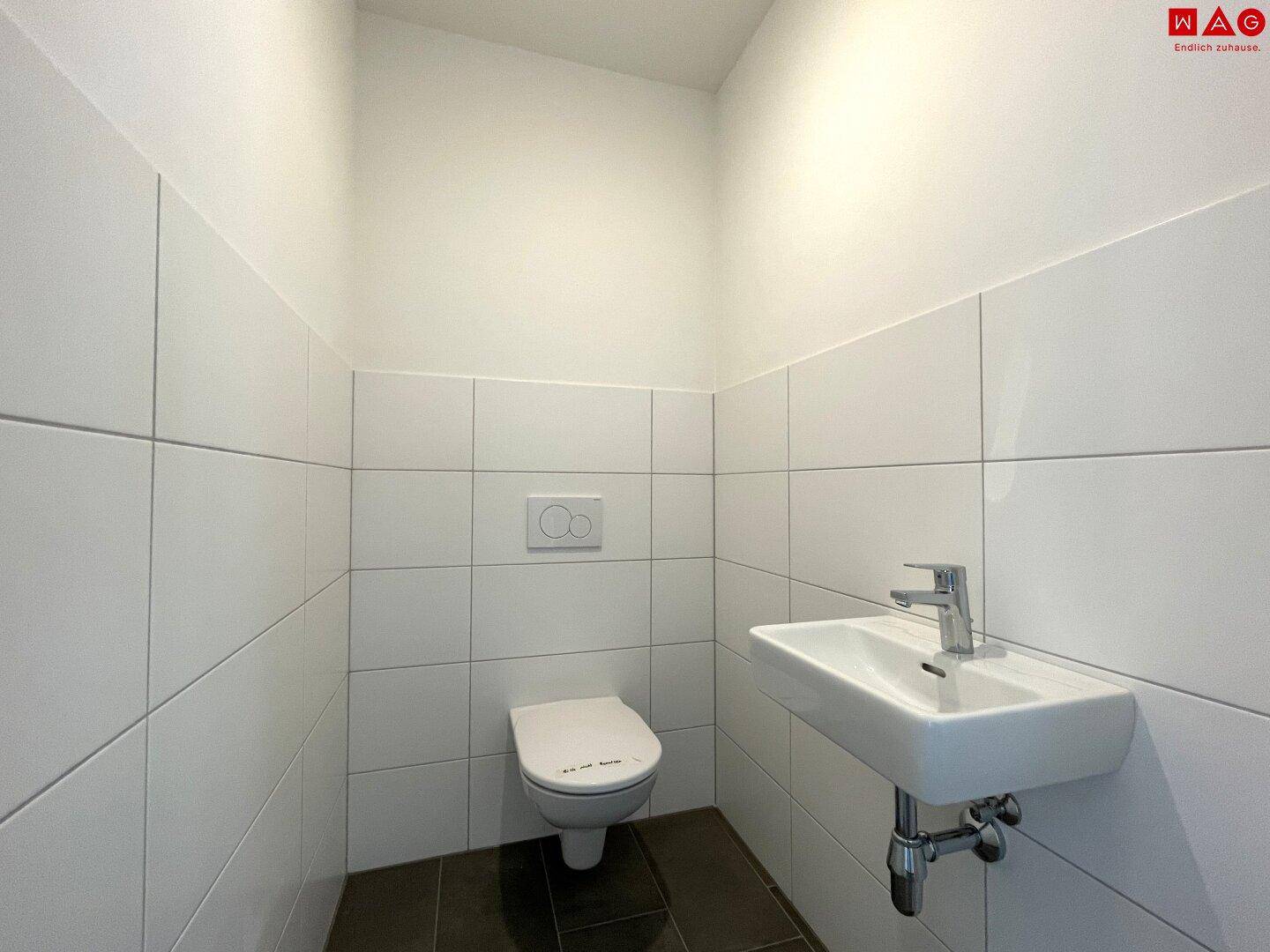 Modernes Gäste-WC mit wandhängender Toilette, kleinem Waschbecken und weißen Fliesen.