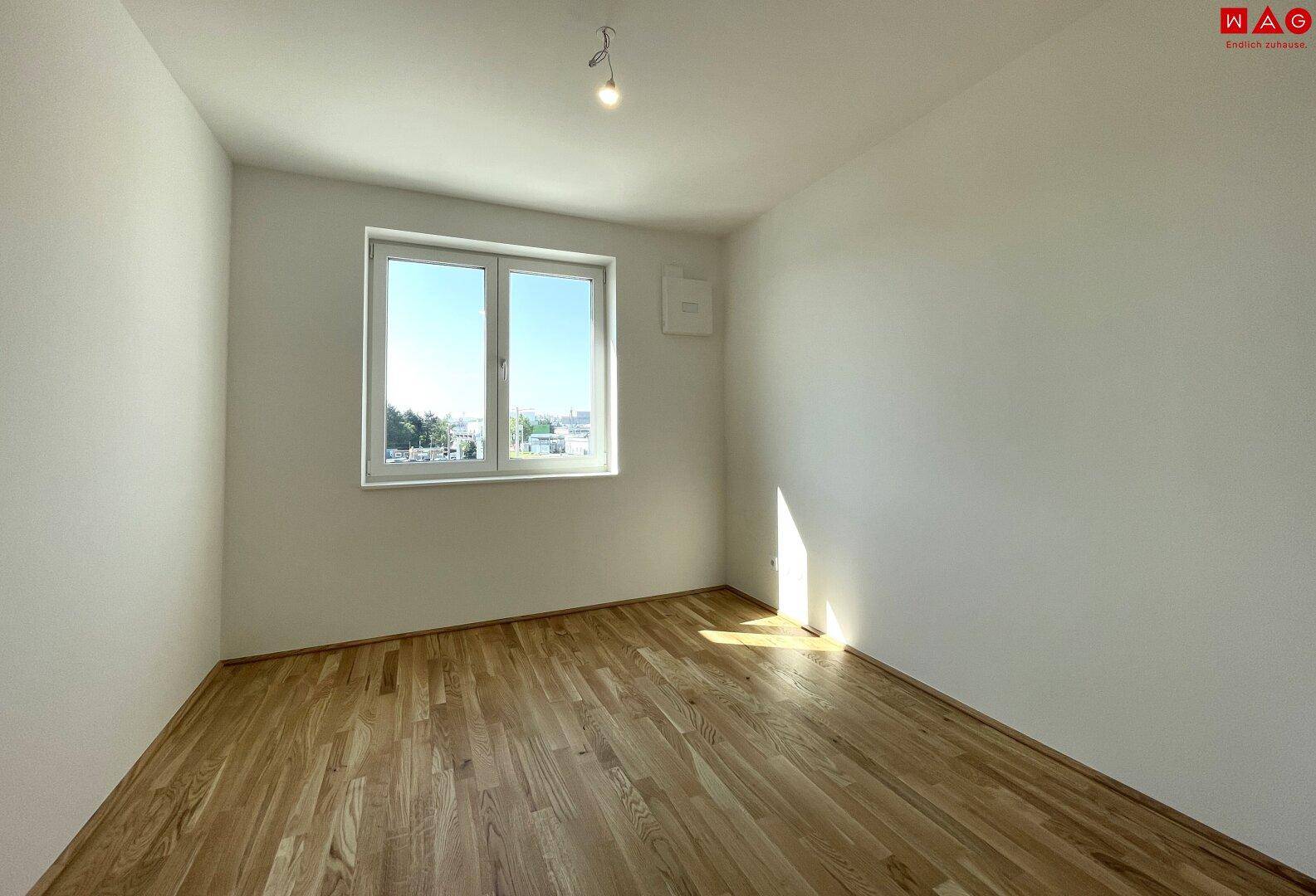Leeres Schlafzimmer mit Holzboden und einem großen Fenster, das viel Licht hereinlässt.