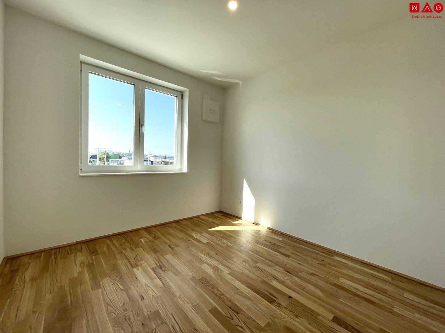 Leeres Schlafzimmer mit Holzboden und einem großen Fenster, das viel Licht hereinlässt.