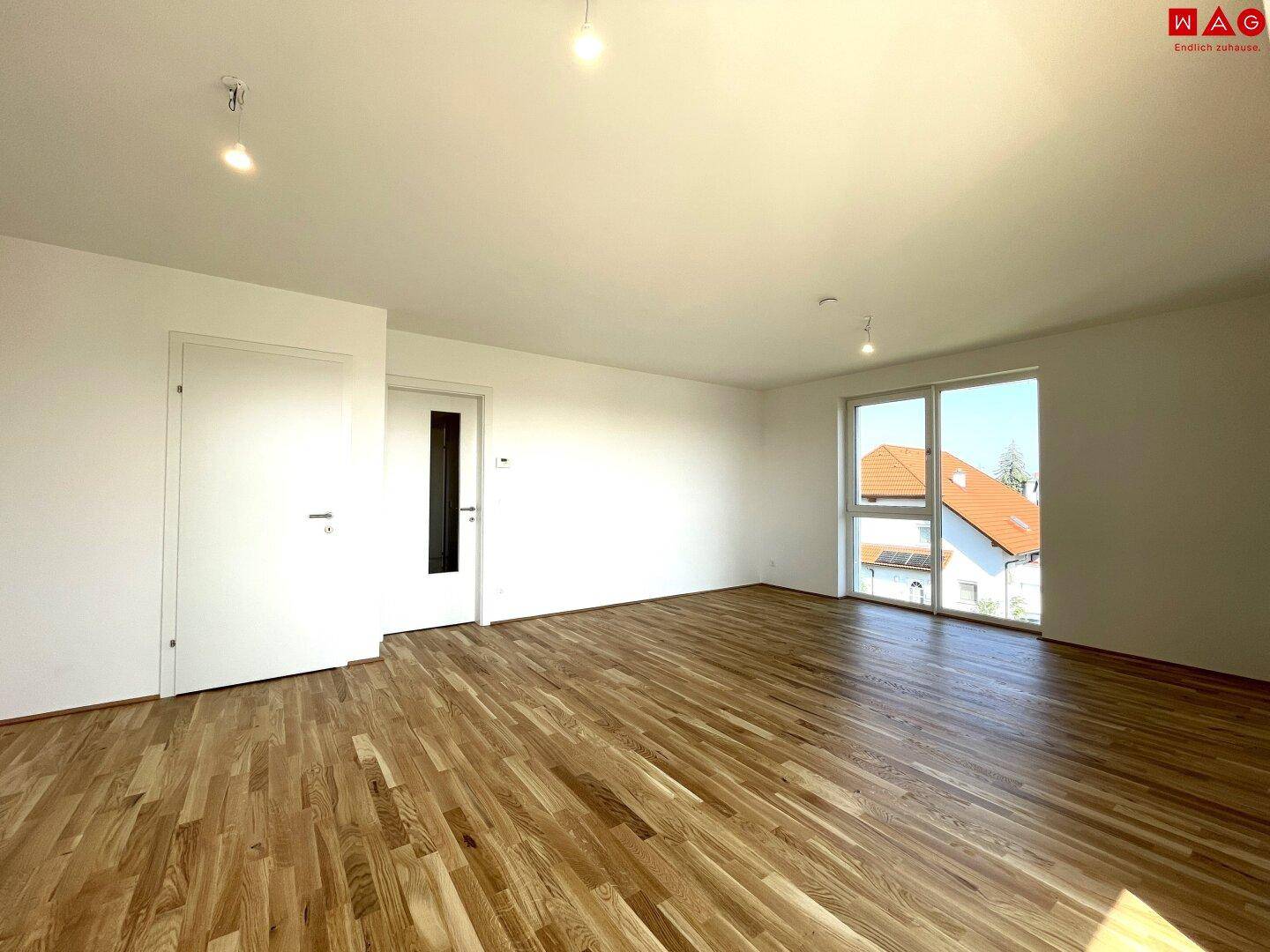 Geräumiges Wohnzimmer mit Holzboden und großem Fenster, das viel Tageslicht hereinlässt.