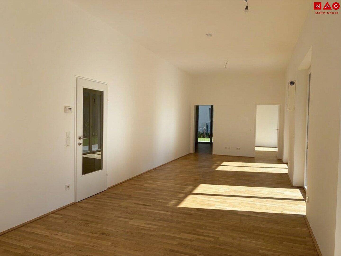 Geräumiges, leeres Wohnzimmer mit Holzboden und viel Tageslicht durch die Fenster.