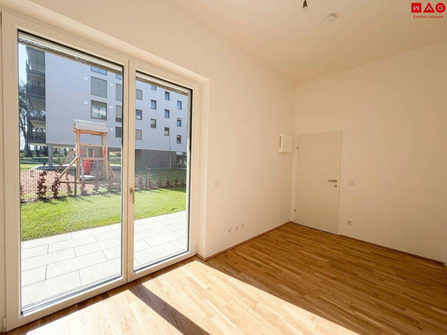 Geräumiges Zimmer mit Holzboden und Blick auf den Spielplatz durch große Fenster.