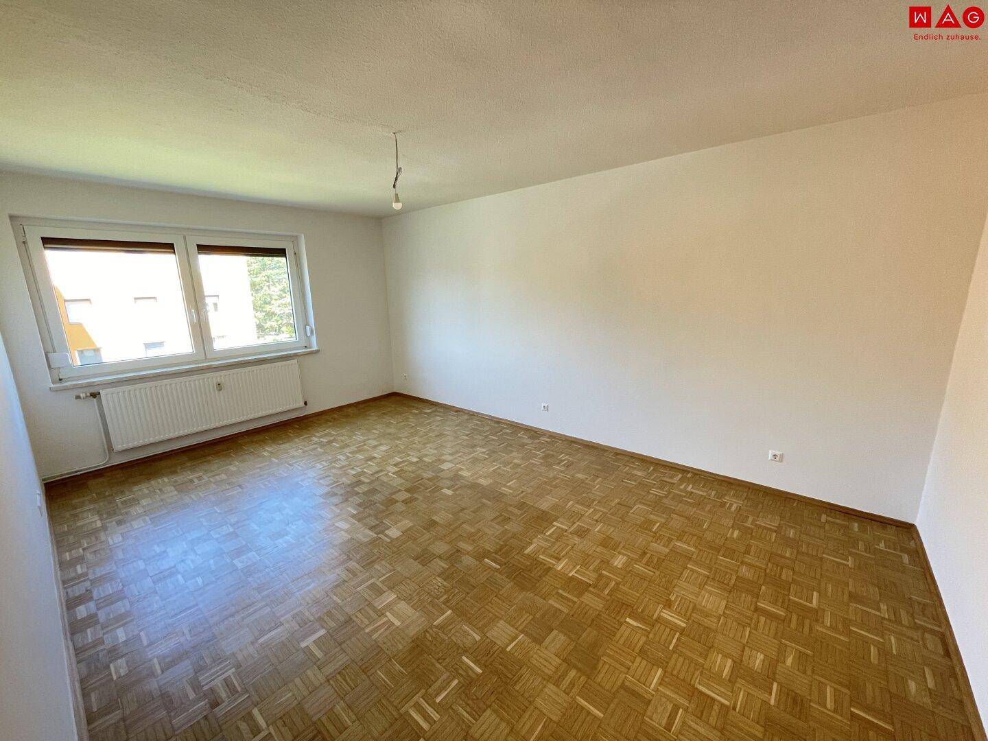 Geräumiger Raum mit klassischem Parkettboden, weißen Wänden und Fenster mit Heizkörper.