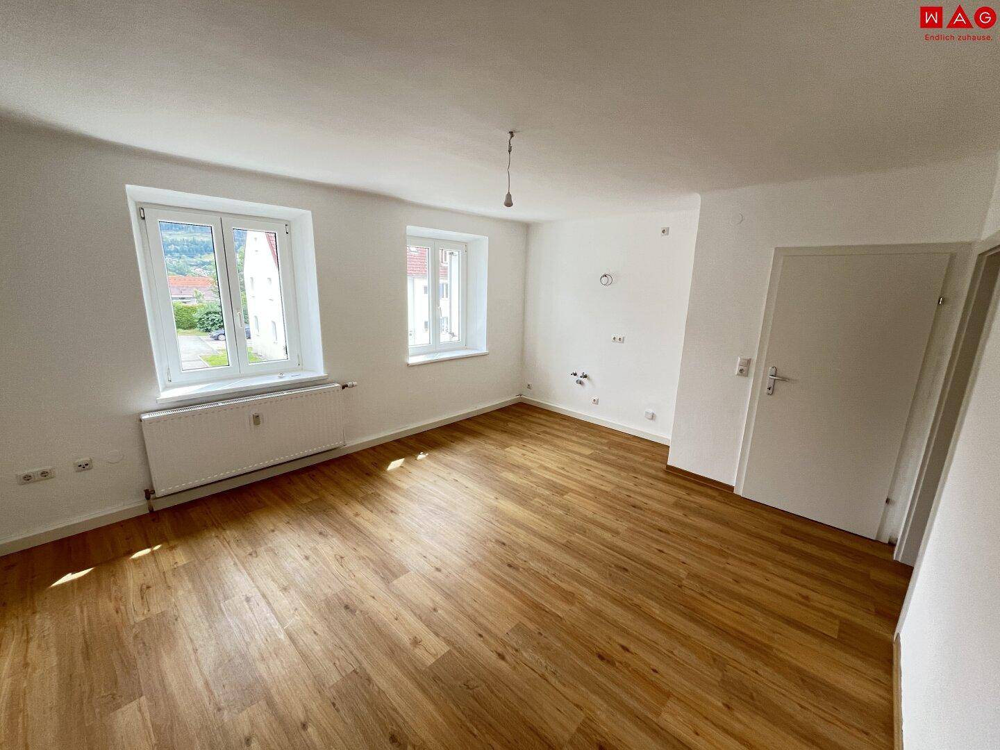 Helles Zimmer mit Laminatboden und zwei Fenstern mit Blick nach draußen.