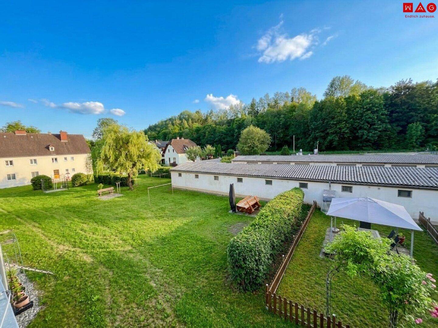 Weitläufige Grünflächen mit Hecken und weiteren Gebäuden unter strahlend blauem Himmel.