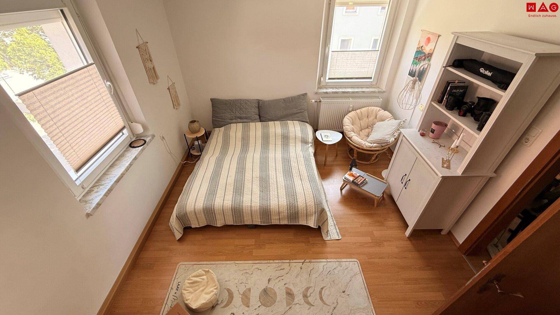 Helles Schlafzimmer mit Holzboden, einem Bett, einem Regal und einem bequemen Sessel am Fenster.