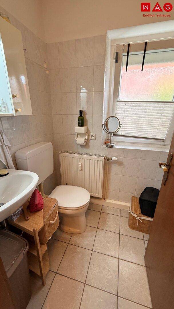 Helles Badezimmer mit WC, Waschbecken, Heizkörper und beigefarbenen Fliesen an Wänden und Boden.