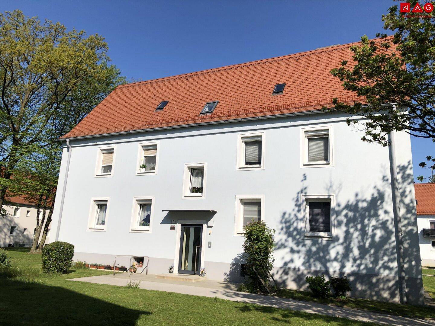 Wohngebäude mit hellblauer Fassade, rotem Satteldach und grüner Rasenfläche im Vordergrund.