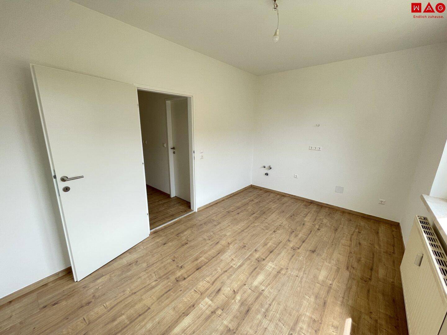 Leerer Raum mit hellem Laminatboden, weißen Wänden und Heizkörper unter dem Fenster.