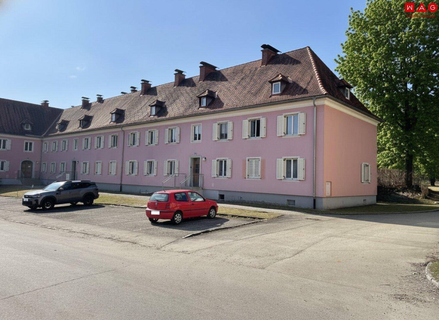 Historisches Gebäude mit rosa Fassade, Satteldach und vielen Dachgauben, umgeben von Grünflächen.