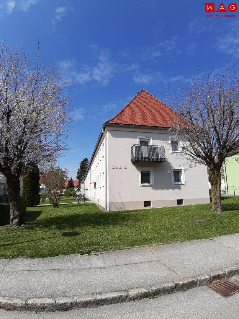 Gebäudeansicht mit rotem Ziegeldach, Balkon und gepflegter Grünfläche unter blauem Himmel.
