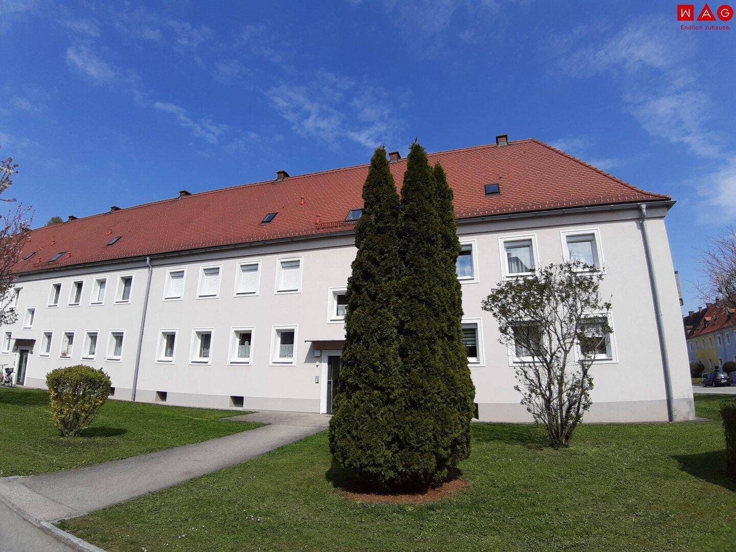 Ansicht eines gepflegten Mehrfamilienhauses mit rotem Ziegeldach und grüner Bepflanzung.