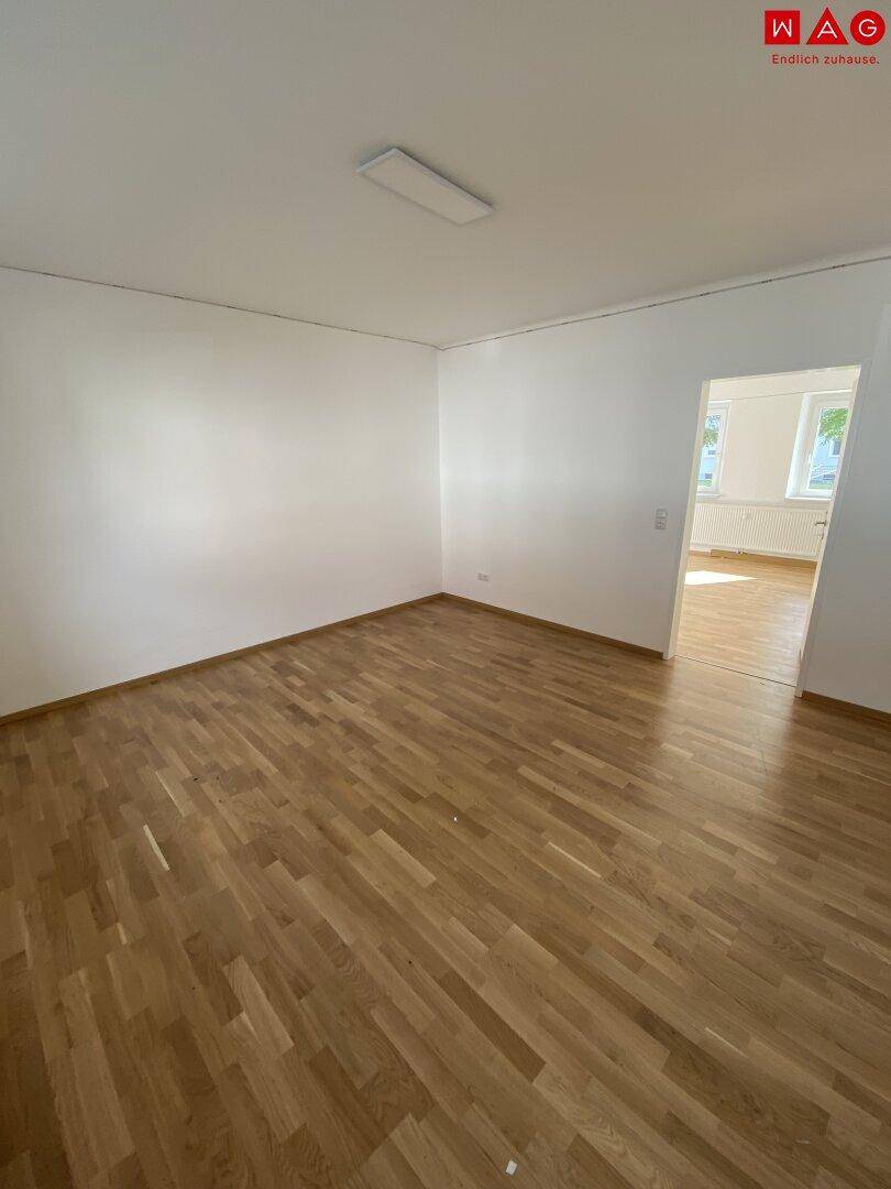 Lichtdurchflutetes Wohnzimmer mit Holzparkettboden und Zugang zu einem weiteren Raum.