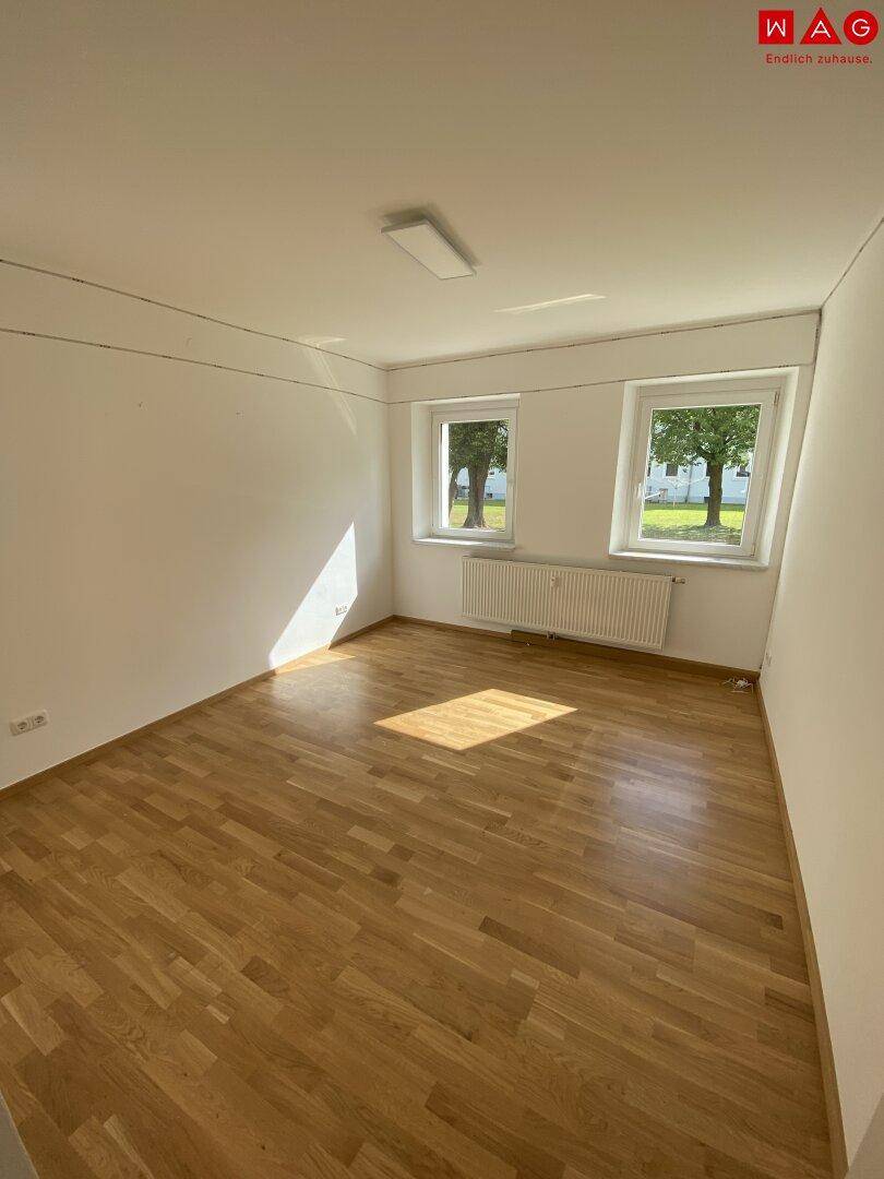 Sonniges Wohnzimmer mit Holzparkettboden und zwei Fenstern, die eine angenehme Atmosphäre schaffen.