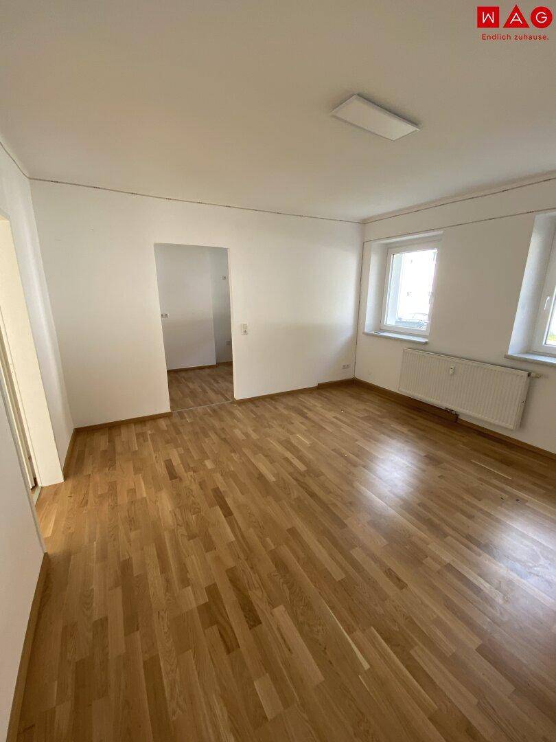 Helles Wohnzimmer mit Holzparkettboden und zwei Fenstern, die viel Tageslicht hereinlassen.