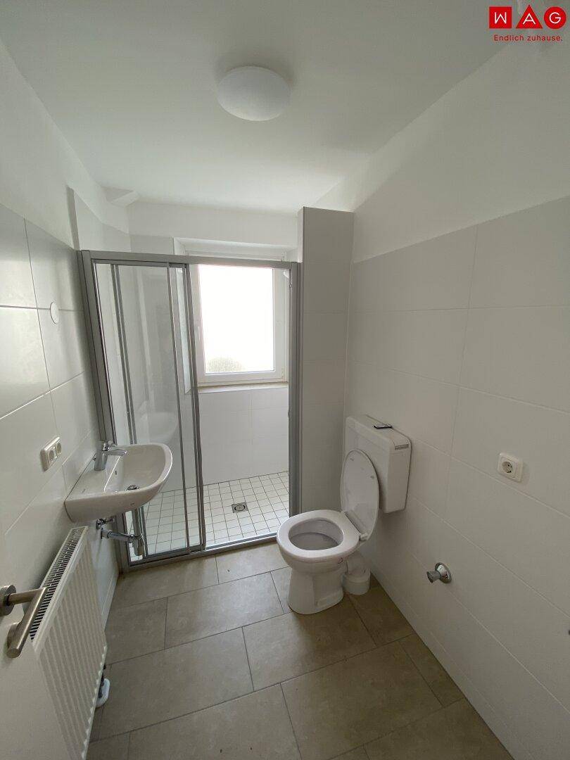 Helles Badezimmer mit Dusche, WC, Waschbecken und Fenster, ausgestattet mit weißen Fliesen.
