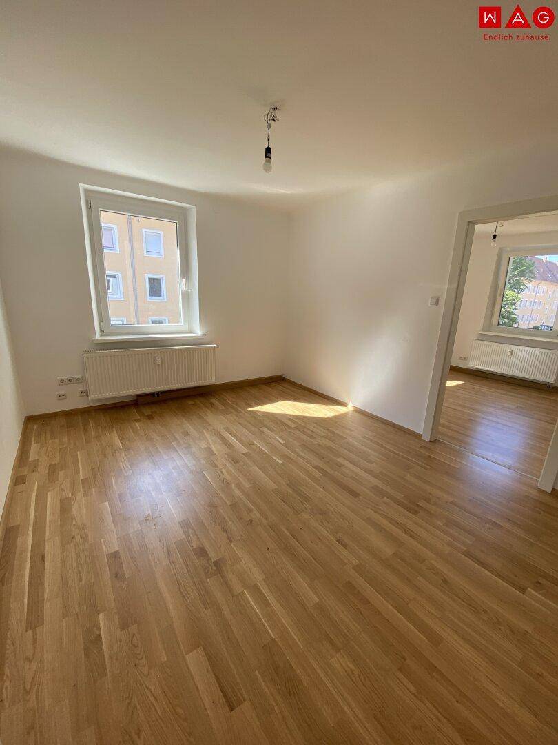 Helles Zimmer mit Parkettboden und Fenster, das viel Tageslicht hereinlässt und einen Ausblick bietet.
