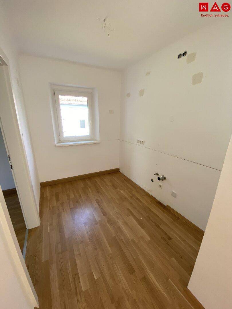 Leerer Küchenraum mit vorbereiteten Anschlüssen und Parkettboden, bereit für die individuelle Kücheninstallation.