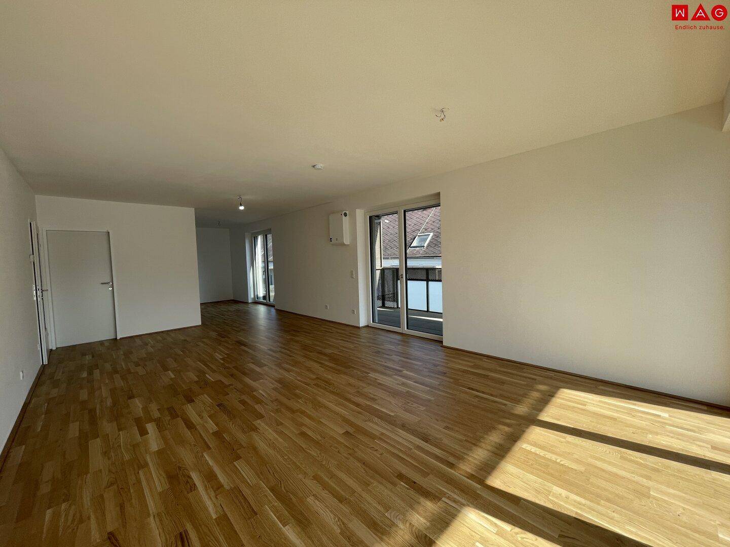 Helles, leeres Wohnzimmer mit Holzboden und Zugang zu einem Balkon oder einer Terrasse.