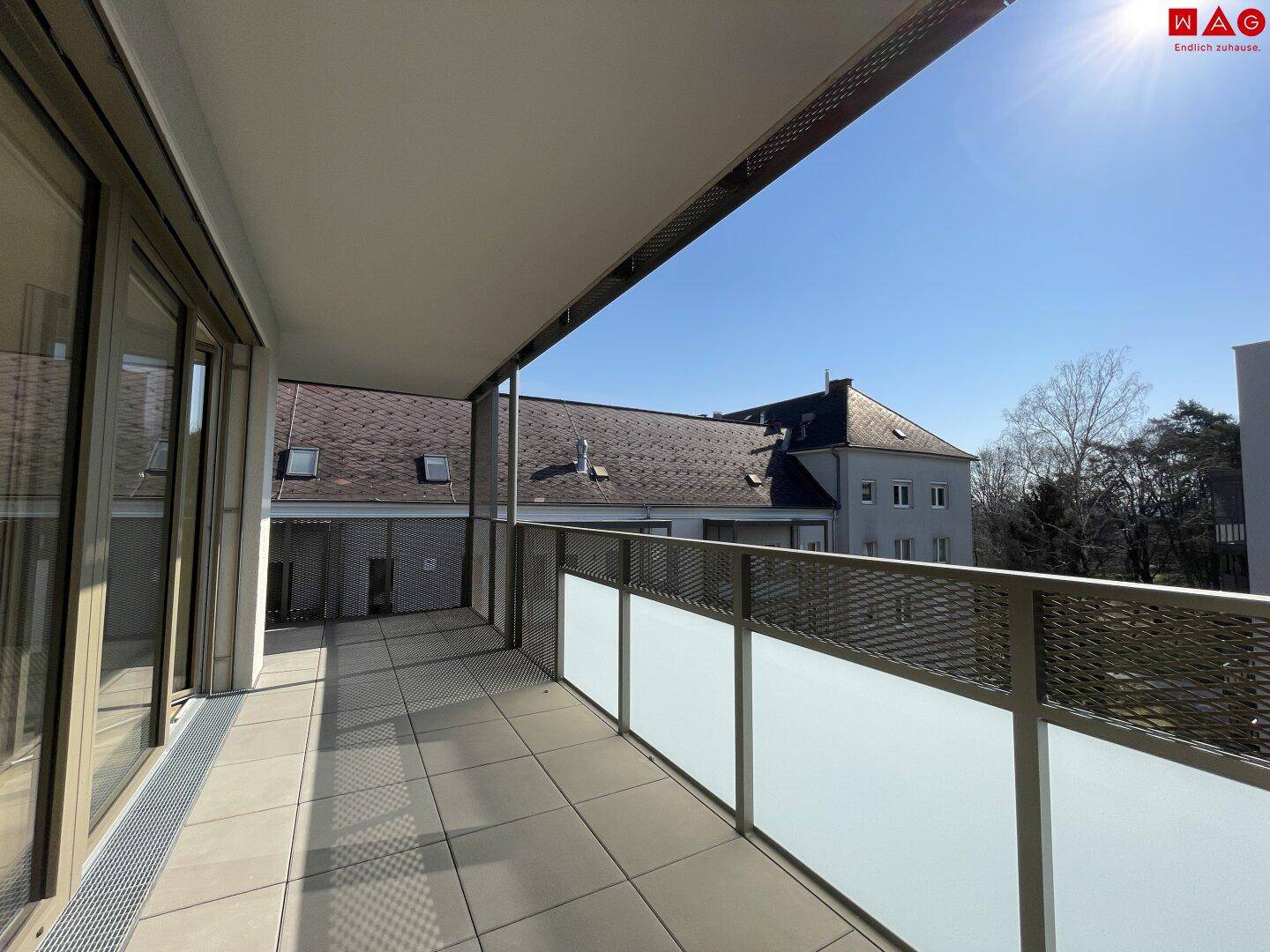 Langer Balkon mit Fliesenboden und Blick auf die umliegenden Gebäude und Bäume.