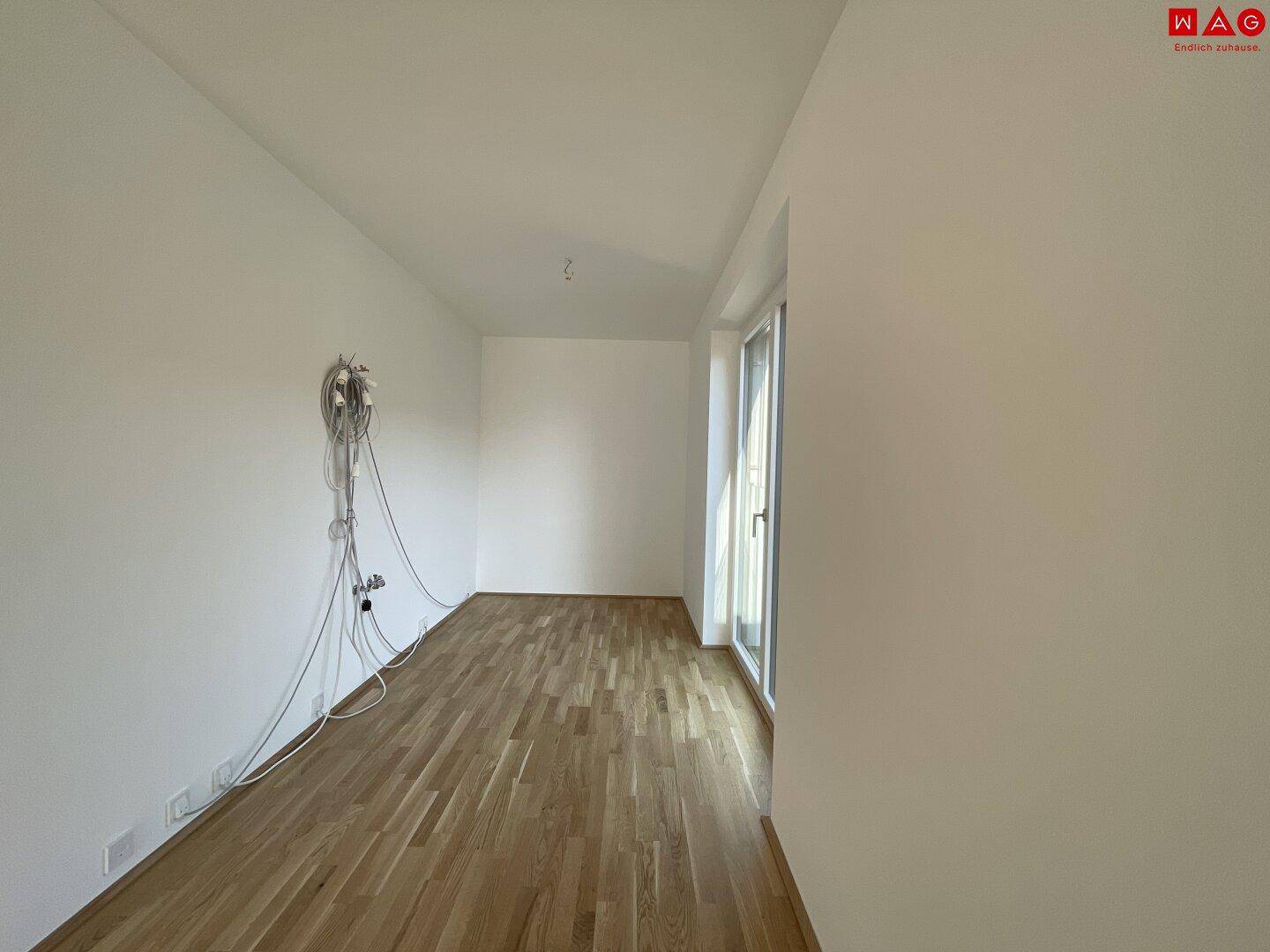 Leeres Schlafzimmer mit Holzboden und großem Fenster, das viel Tageslicht hereinlässt.