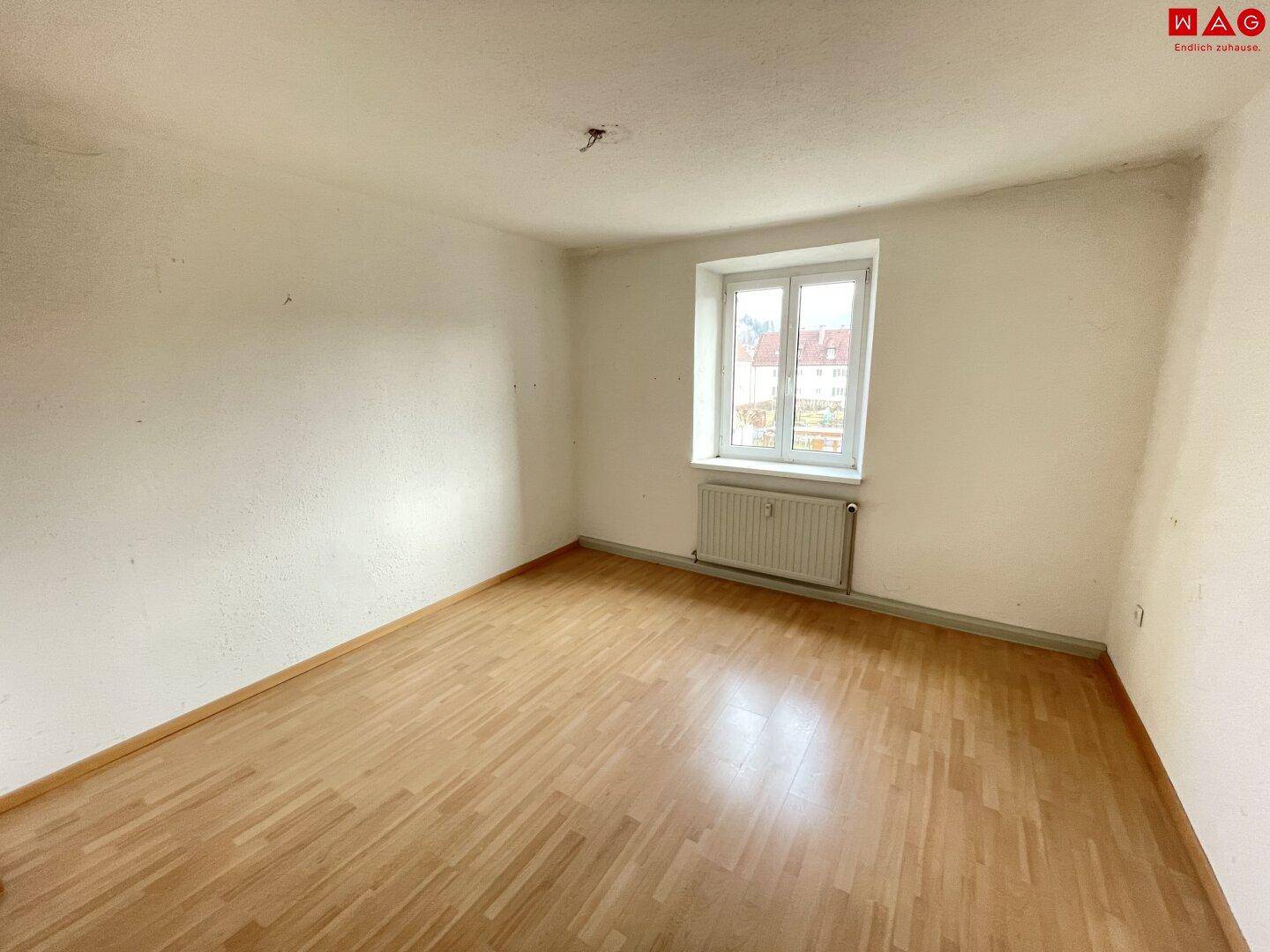 Geräumiger, leerer Raum mit Holzparkett und Fenster, das viel Tageslicht hereinlässt.