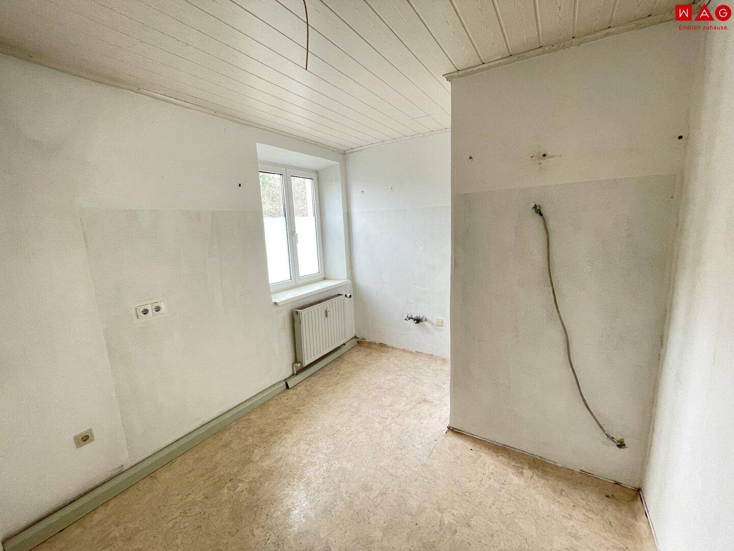 Leerer Raum mit Fenster, Heizkörper und einfacher Holzdeckenverkleidung, bereit zur Renovierung.