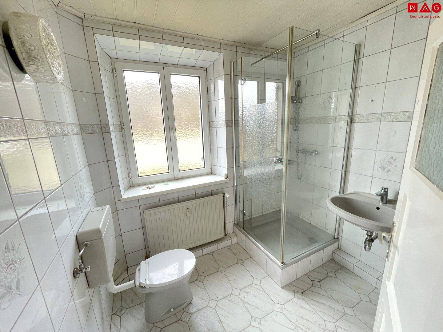 Helles Badezimmer mit Duschkabine, WC und Waschbecken, gefliest in Weiß und mit Fenster.