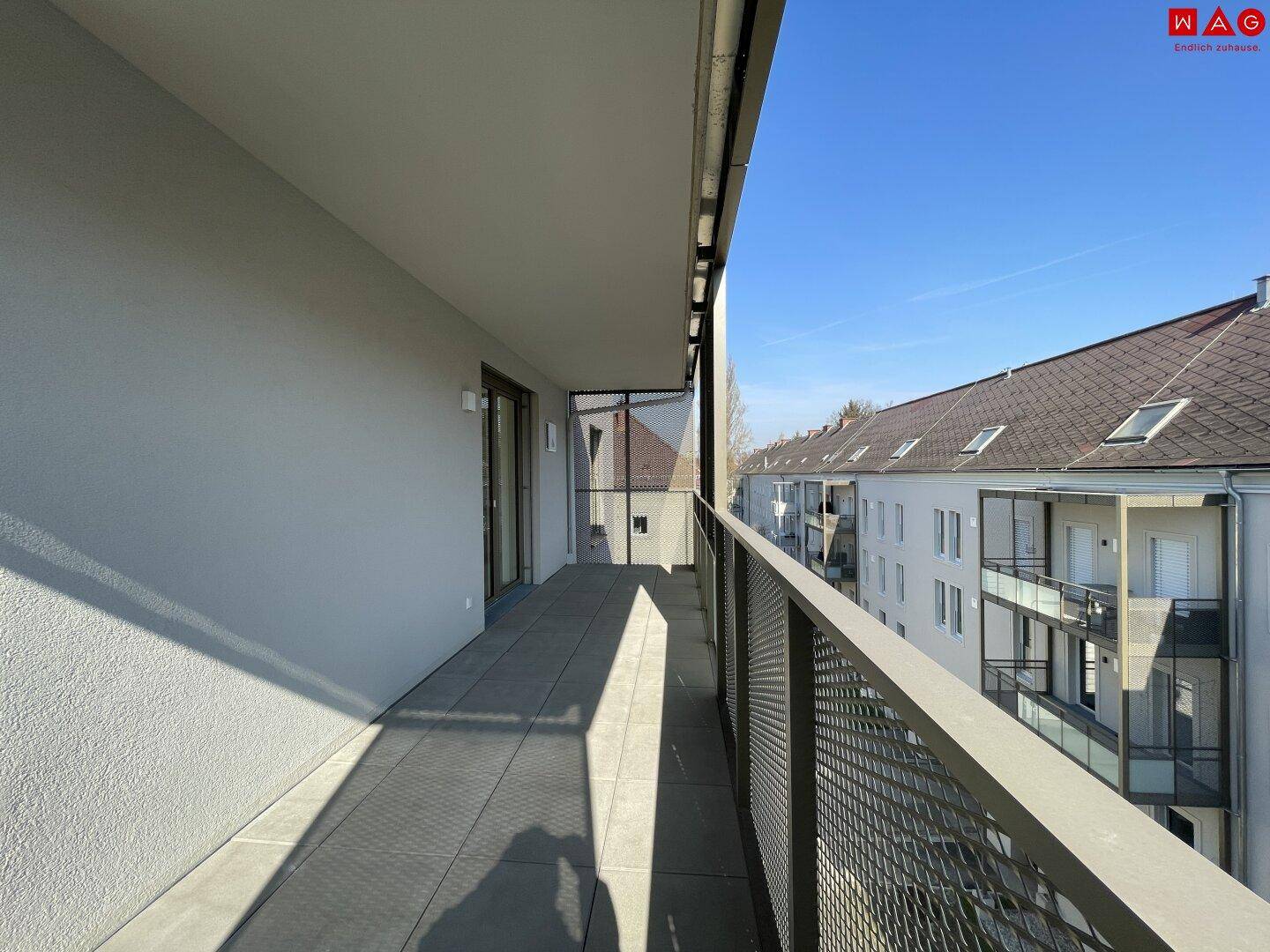 Langer, leerer Balkon mit Fliesenboden und Metallgeländer, bietet Blick auf umliegende Gebäude.