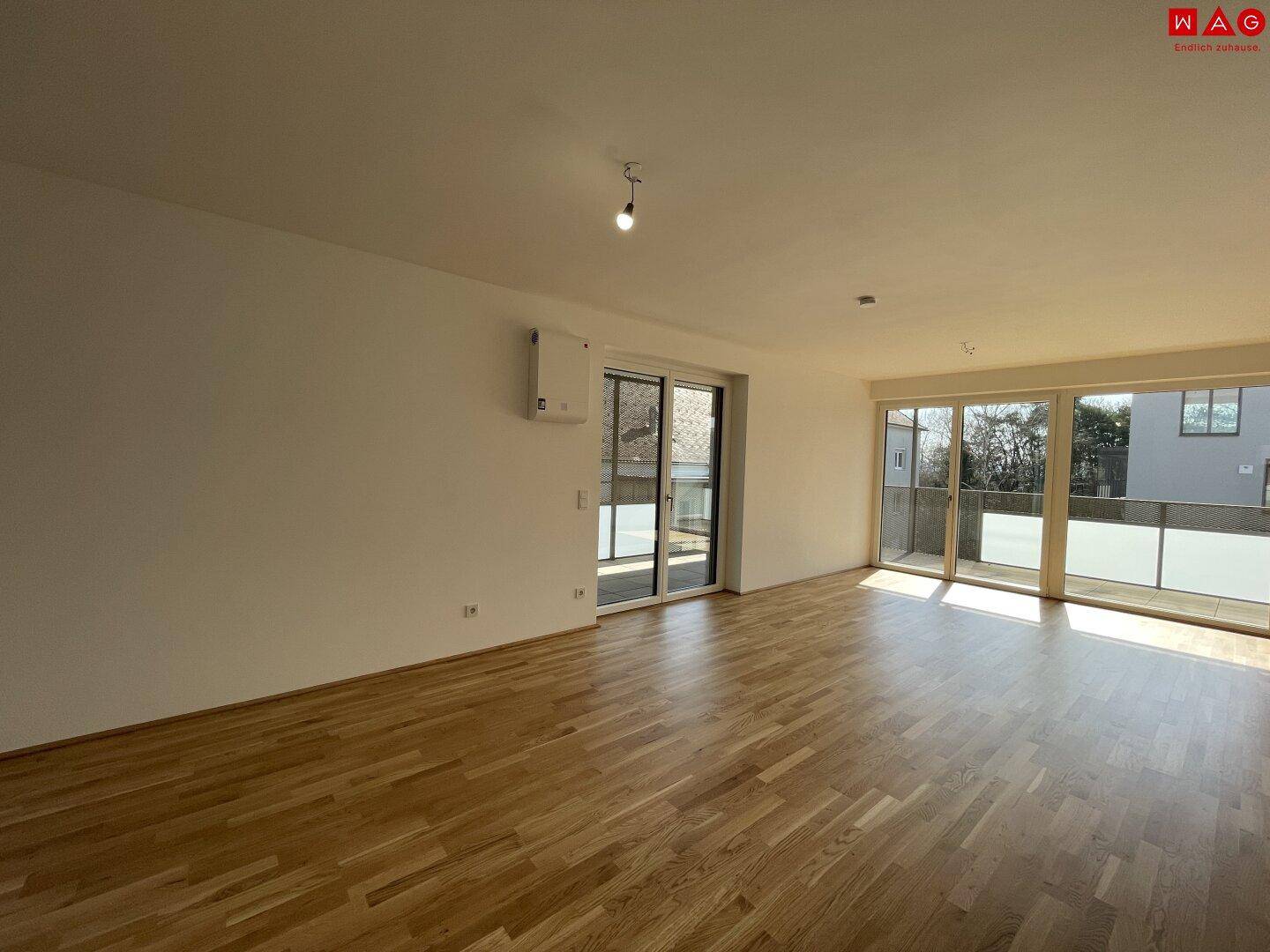 Heller, leerer Wohn-Essbereich mit Holzboden, großen Fenstern und direktem Zugang zum Balkon.