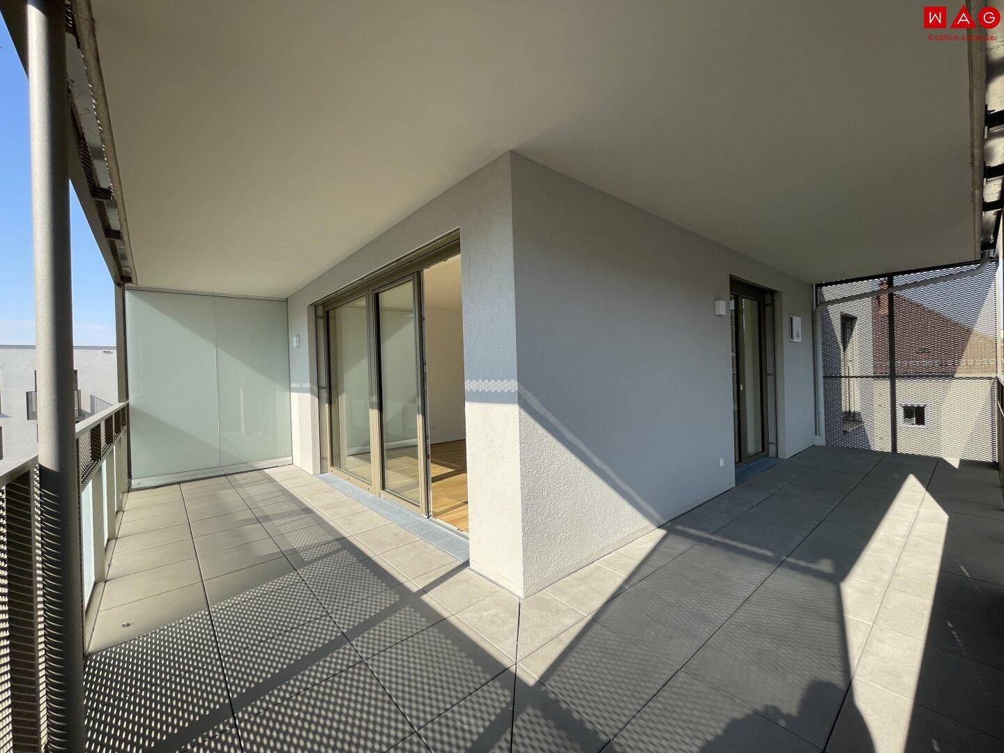 Heller, leerer Balkon mit Fliesenboden und moderner Glasbrüstung unter blauem Himmel.