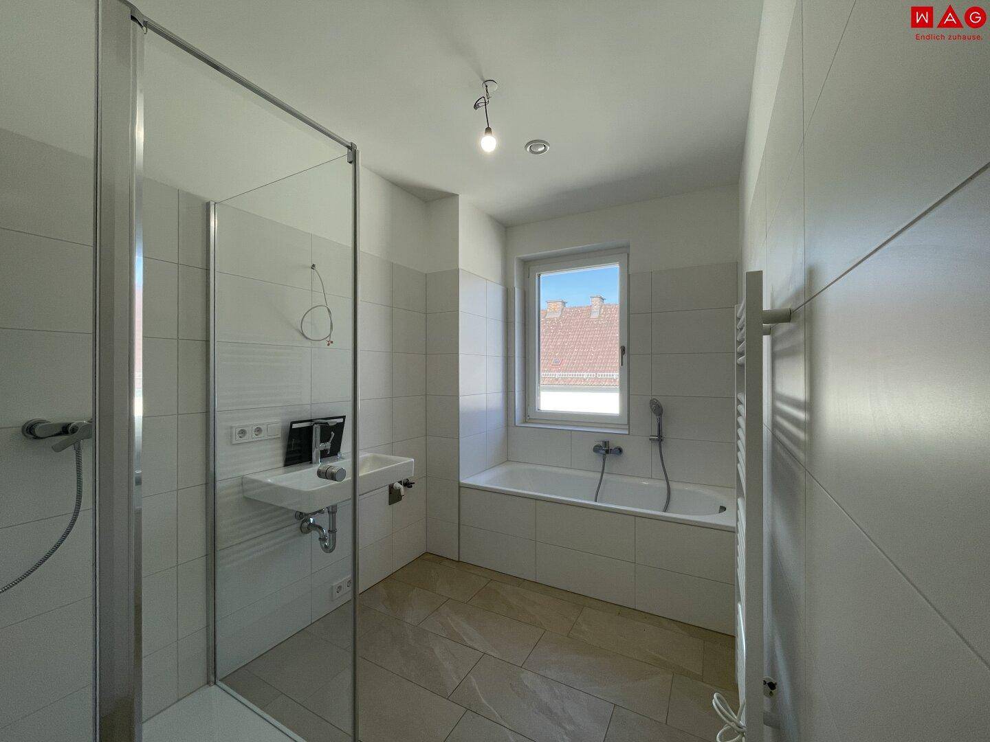 Helles, leeres Badezimmer mit Duschkabine, Badewanne, kleinem Waschbecken und Fenster.