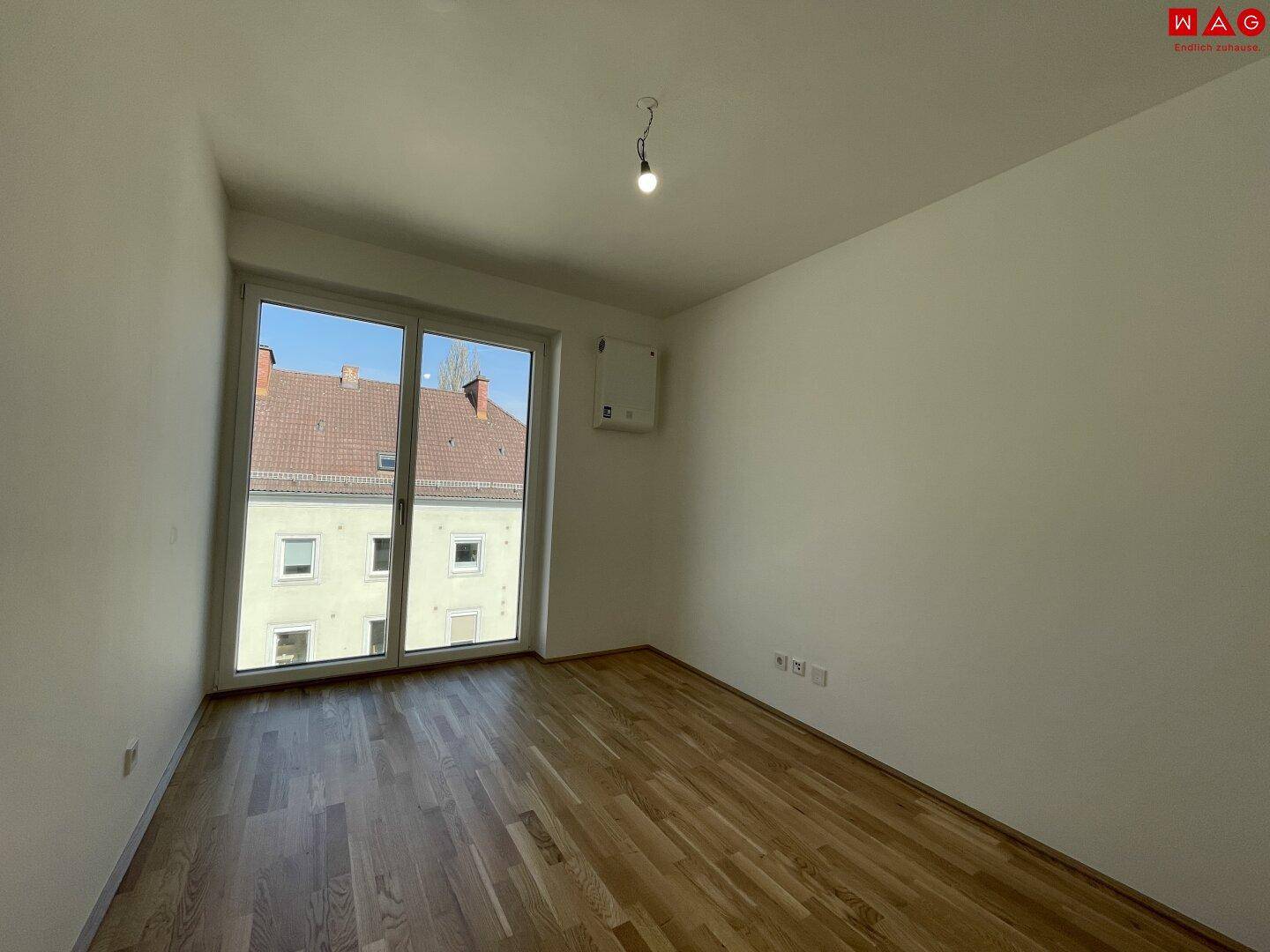 Heller, leerer Raum mit Holzboden und großem Fenster, ideal als Schlaf- oder Arbeitszimmer.