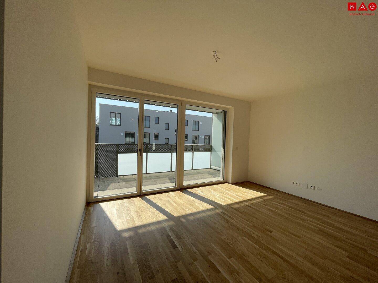 Helles, leeres Schlafzimmer mit Holzboden und großen Fenstern zum Balkon.