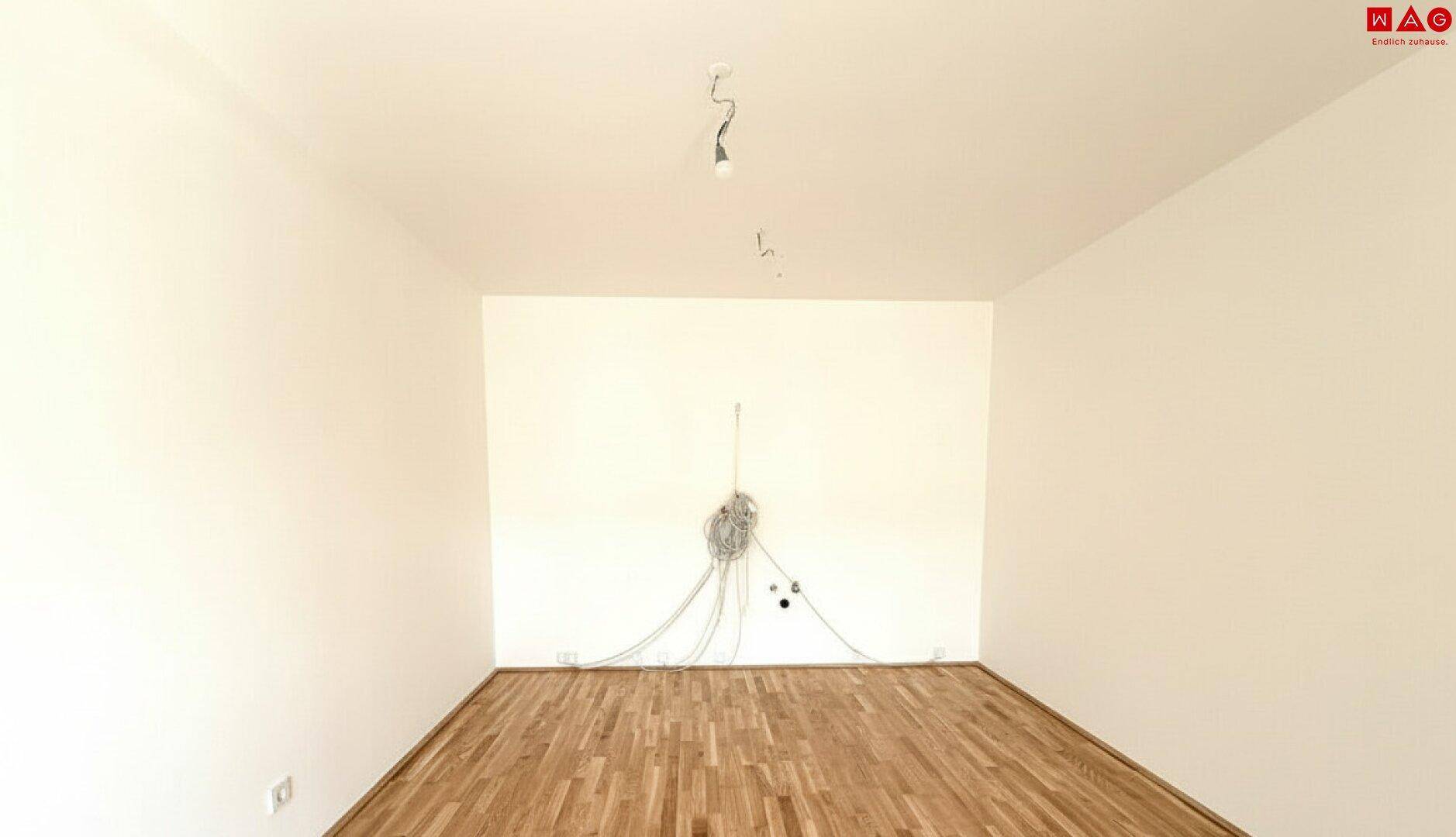 Leerer Raum mit Parkettboden und vorbereiteten Anschlüssen für Beleuchtung und Medien.