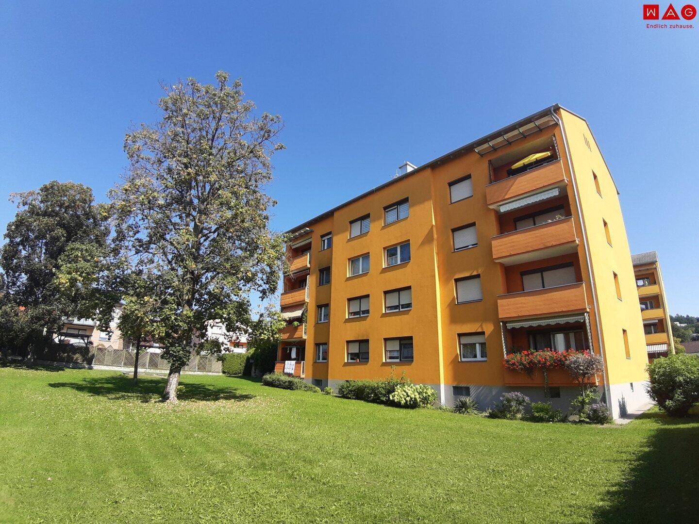 Sonnenbeschienenes Mehrfamilienhaus mit gelber Fassade, Balkonen und gepflegten Grünflächen bei blauem Himmel.