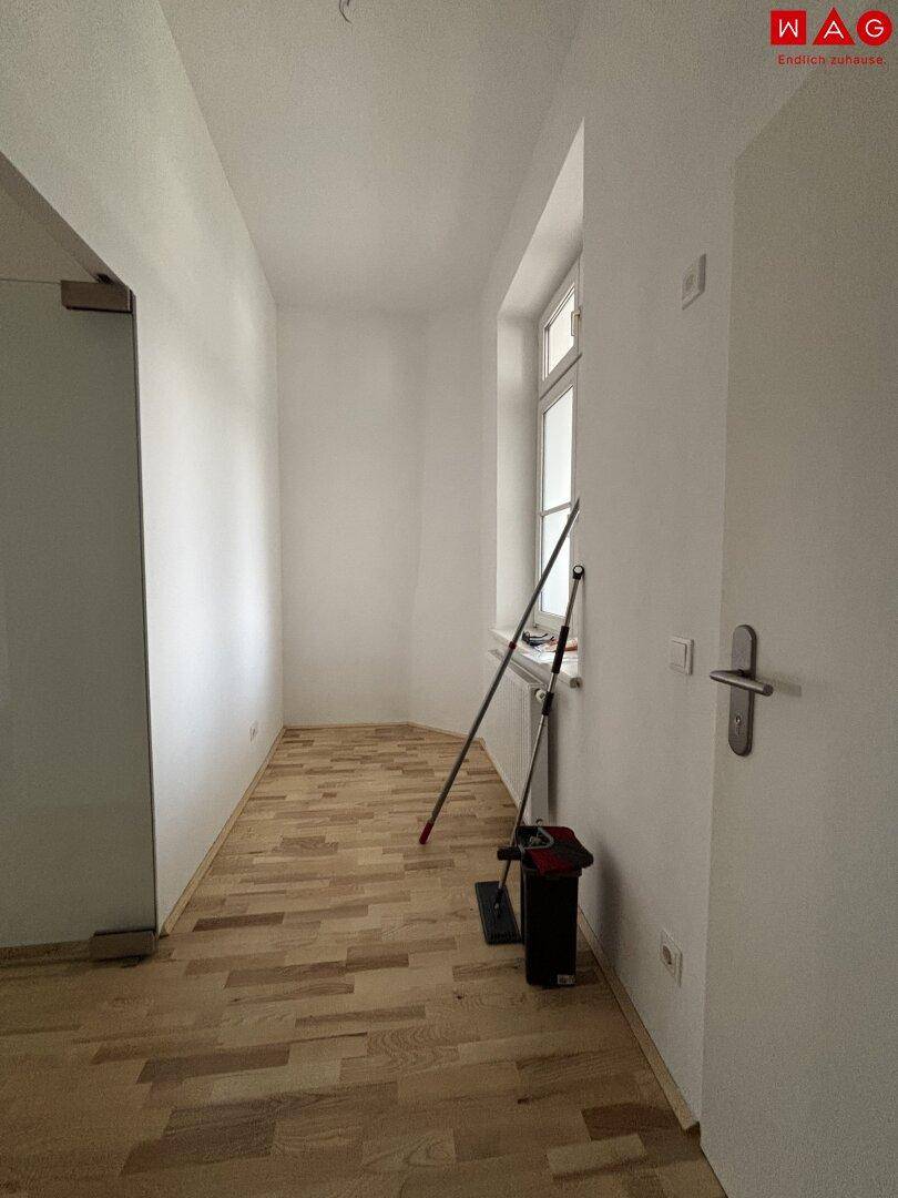 Leerer Raum mit hellem Parkettboden und einem Fenster, das Tageslicht hereinlässt.