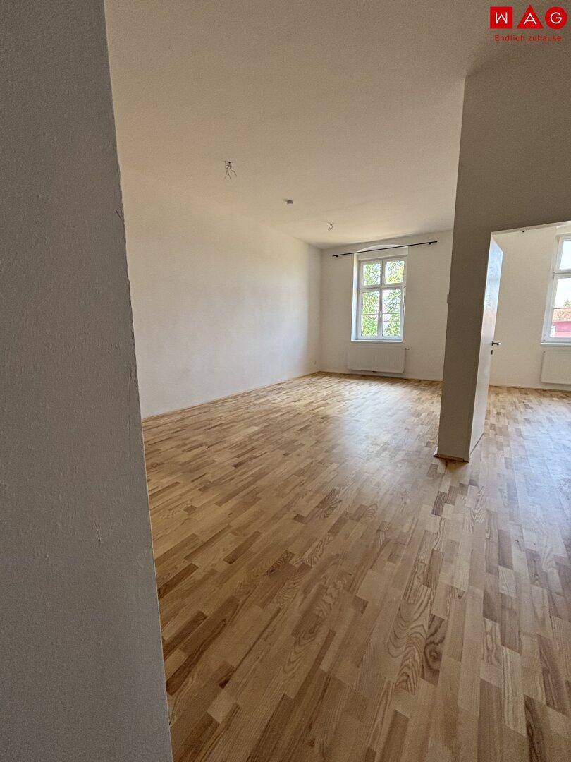 Großzügiger, leerer Wohnraum mit hellem Parkettboden und mehreren Fenstern für viel Tageslicht.