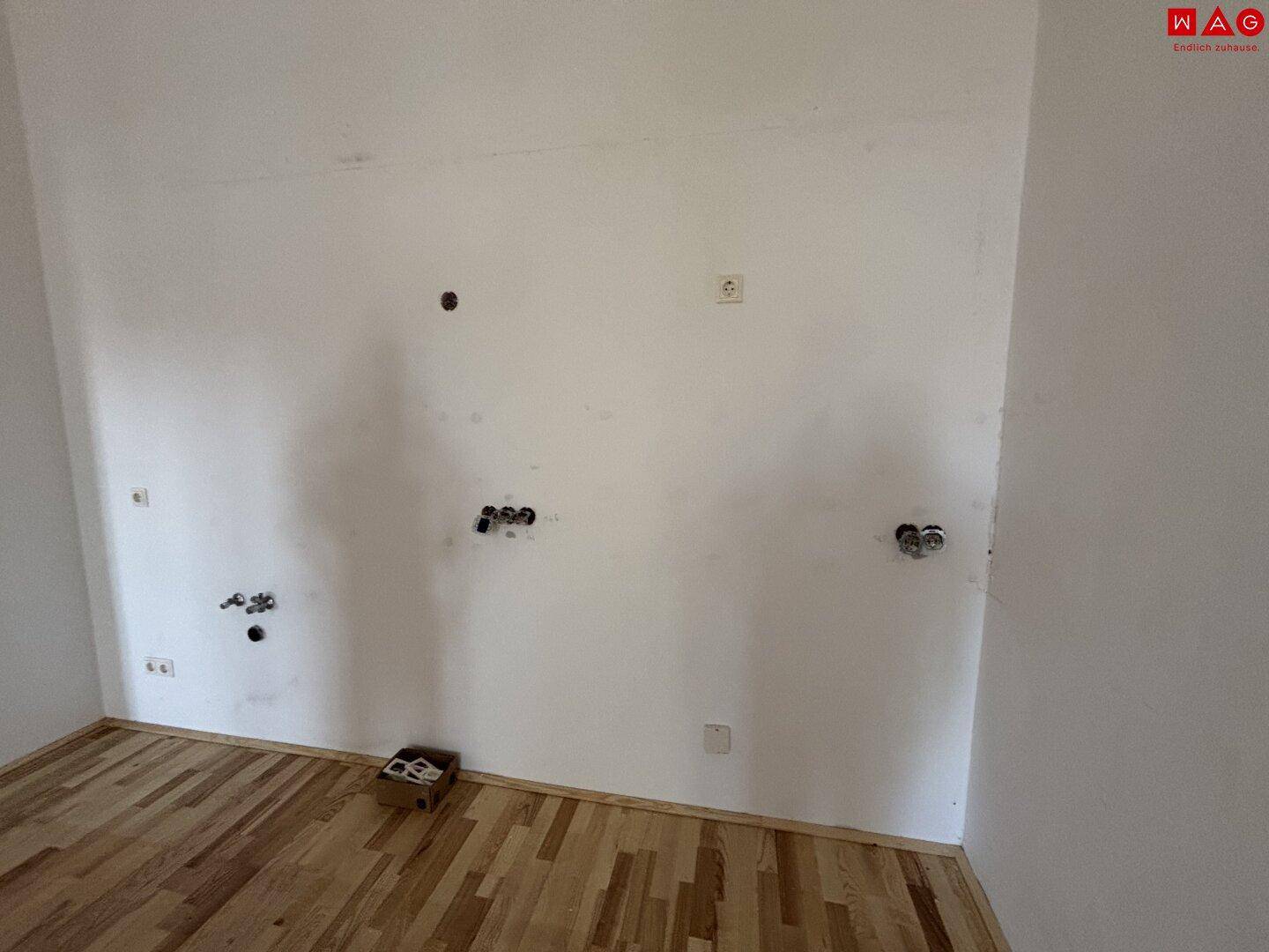 Leerer Küchenbereich mit vorbereiteten Anschlüssen und hellem Parkettboden, bereit für die Installation.