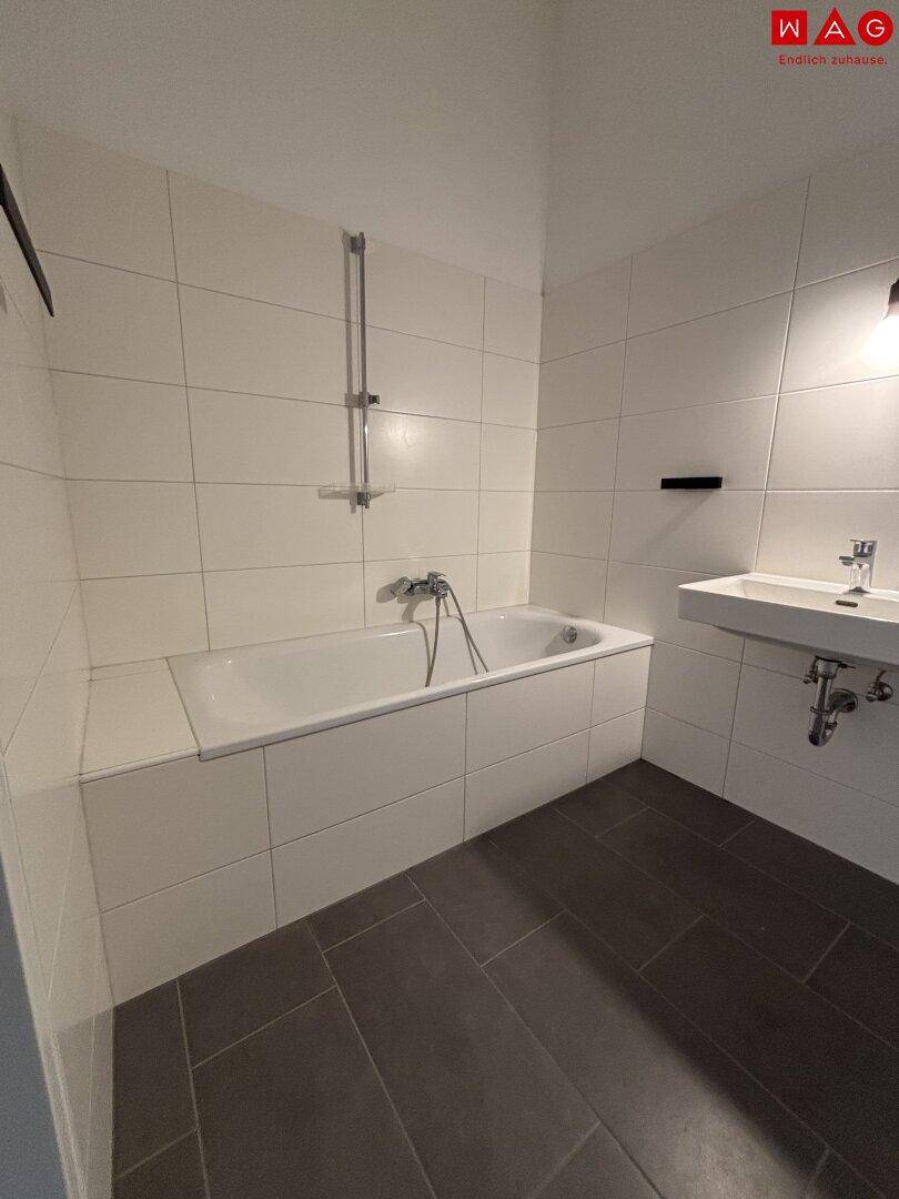 Modernes Badezimmer mit Badewanne, Waschbecken und dunklen Bodenfliesen in schlichtem Design.